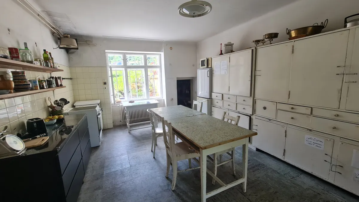 A large kitchen with built-in white cupboards, a stone floor, a long wooden table and a window onto green trees.