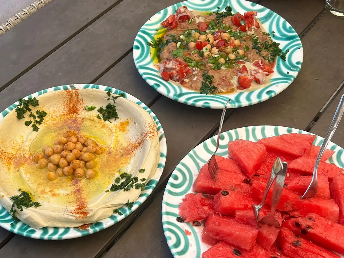 A garden table with two plates of hummus, fatteh and a plate of cut watermelon.