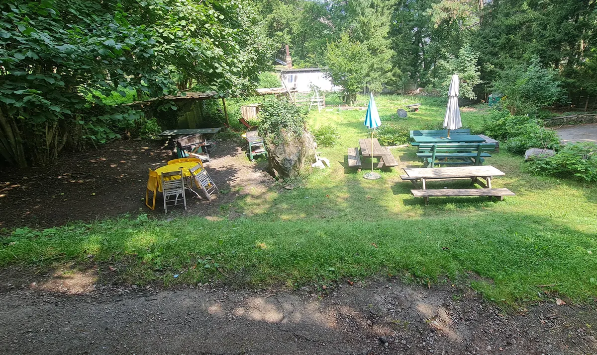 The garden at midday: green picnic tables, folded umbrellas, a yellow table under a hazel tree and a mossy boulder.