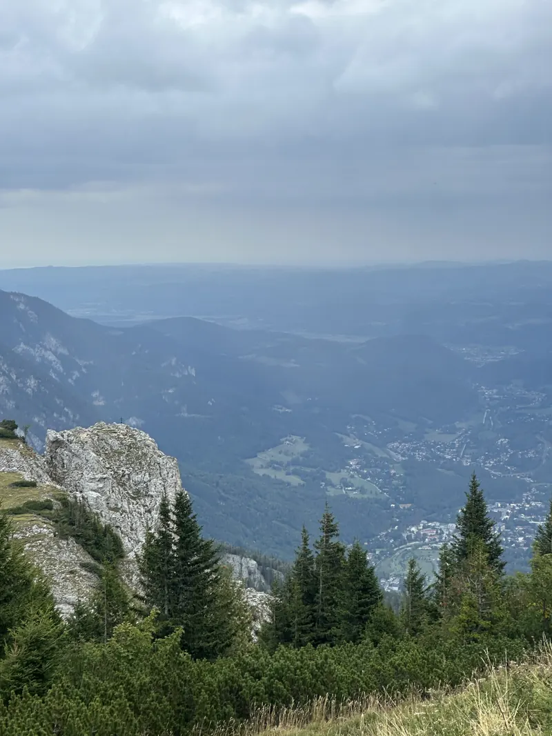 The view from the top of the Rax: pine trees, a limestone cliff and the valley far below under cloud.