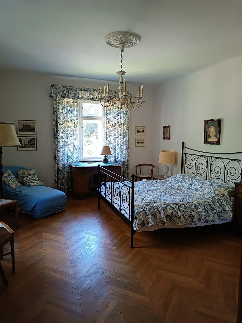 An iron bed with a flowered cover under a ceiling rose and chandelier, blue floral curtains behind.