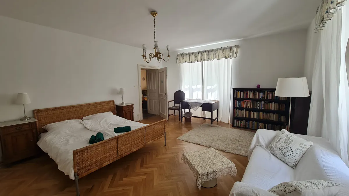 A large room with a double bed, a bookcase full of books, a writing desk and a white sofa.