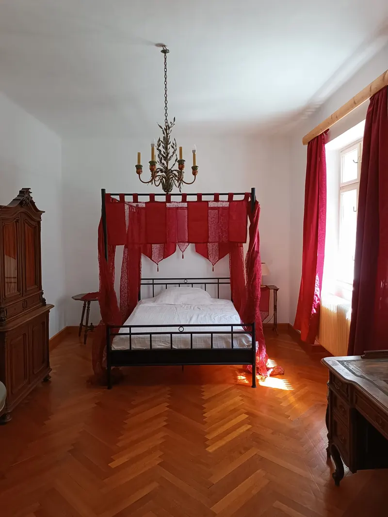 An iron four-poster bed hung with red cloth under a chandelier, between two carved wardrobes.