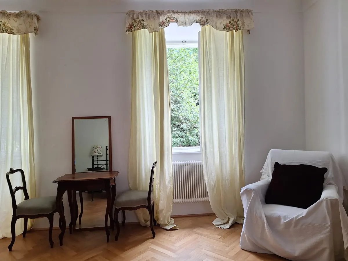 Two tall windows with cream curtains, a small table with a mirror and chairs, and an armchair under a white cover.